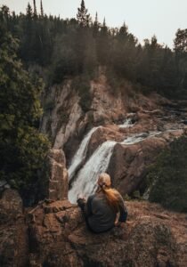 Back View Photo of a Woman Sitting on Rocks Overlooking the Waterfalls